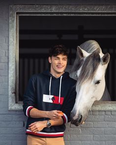 a young man standing next to a white and gray horse in an open window with his arm around the horse's head