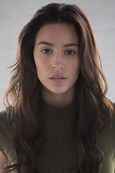 a woman with long brown hair wearing a green shirt and looking at the camera while standing in front of a white wall