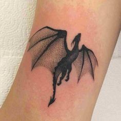 a small black dragon tattoo on the ankle, with its wings spread out and it is flying