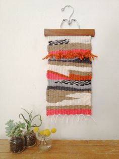 a woven wall hanging with orange and pink accents on a wooden shelf next to two vases