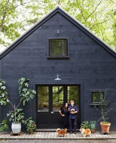 a man and woman standing in front of a black house with two dogs on the porch