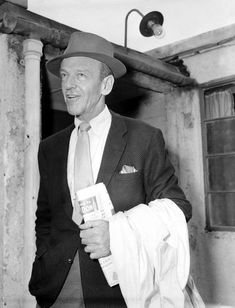 an old man wearing a suit and tie holding a book in his hand while standing next to a building