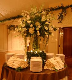 a table topped with lots of plates covered in white flowers and greenery next to a tall vase filled with white roses