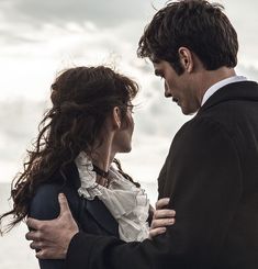 a man and woman standing next to each other in front of the ocean on a cloudy day