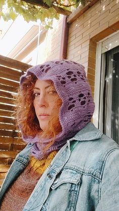 a woman with red hair wearing a purple crocheted hat and jacket standing in front of a window