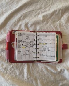 a red planner with notes and pen on top of white sheeted paper that is laying on a bed