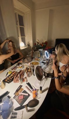 three women sitting at a table with makeup and other items on it, all looking in the same direction