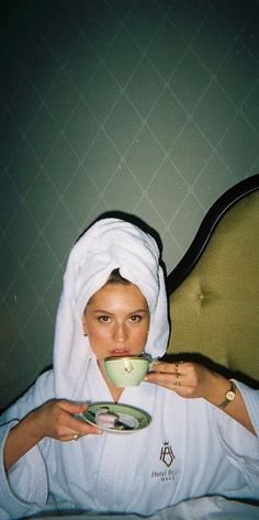 a woman sitting on a bed drinking from a cup and saucer in front of her face