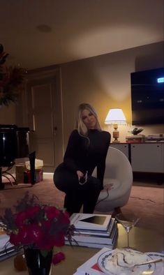 a woman sitting on a chair in a living room next to a table with books