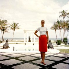 a woman is standing in front of a pool with palm trees and statues behind her