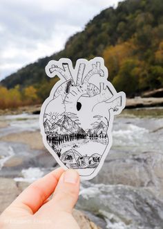 a hand holding up a sticker with a river and mountains in the back ground