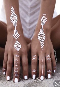 a woman's hands with white painted designs on their fingers and arms, sitting in the sand