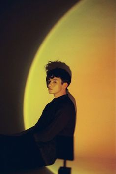 a young man sitting on top of a chair in front of a yellow wall with the sun behind him