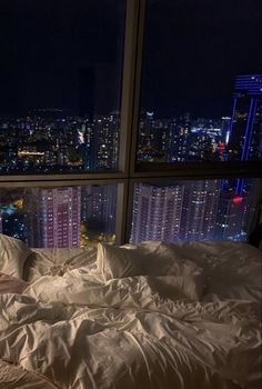 an unmade bed in front of a large window with city lights outside at night