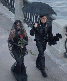 two people dressed in black walking down the street with an umbrella over their heads and one holding a rose