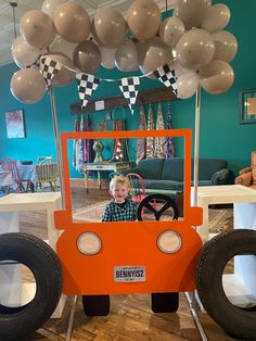 a young boy is sitting in an orange car made out of tires with balloons hanging from the ceiling