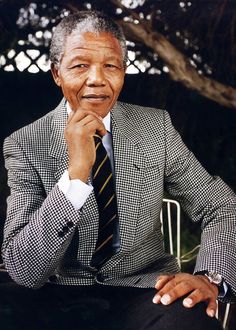 an older man in a suit and tie sitting on a chair with his hand under his chin