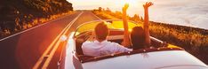 two people in a convertible driving down the road with their hands up to the sky