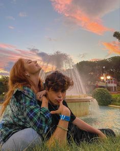 two people sitting in front of a fountain with their arms around each other and looking up at the sky