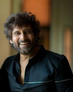 a man with long hair smiling and wearing a black shirt in front of a wall