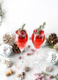 two wine glasses filled with red liquid surrounded by pine cones and christmas decorations on a white surface