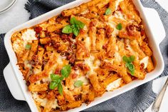 a white casserole dish filled with pasta and meat covered in sauce, cheese and basil