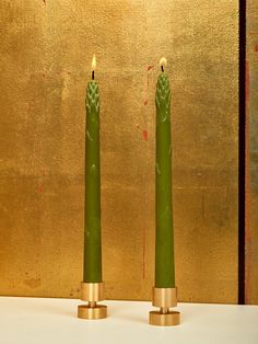 two green candles sitting next to each other on a white counter top in front of a gold wall