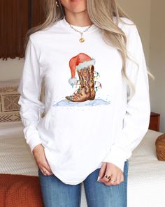 Western Christmas shirt. This long sleeve t-shirt is perfect for the holidays! It features a pair of cowboy boots sitting on snow with a Santa hat on top. The boots are wrapped in colorful Christmas lights. Get into the festive spirit with this fun and unique design!  Made with 100% ring-spun cotton, these personalized long-sleeve shirts come packed with softness and style. Each tee features garment-dyed fabric and comes with a relaxed fit for total comfort in any casual setting. Add your design Christmas Cowboy Boots, Western Christmas Shirts Cricut, Western Christmas Tshirt Ideas, Country Christmas Shirts, Cowboy Boot Tshirt, Christmas Tee Shirts Western, Cowboy Christmas Shirt, Colorful Christmas Lights, Cowgirl Christmas