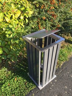 a metal trash can sitting on the side of a road next to bushes and trees