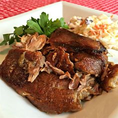 a white plate topped with meat next to coleslaw and carrots on a red tablecloth