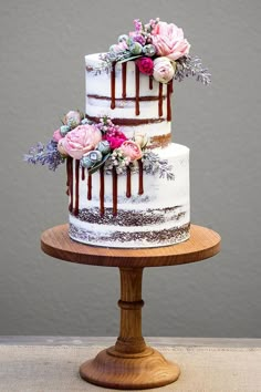 a three tiered cake with flowers and chocolate icing on a wooden stand in front of a gray wall