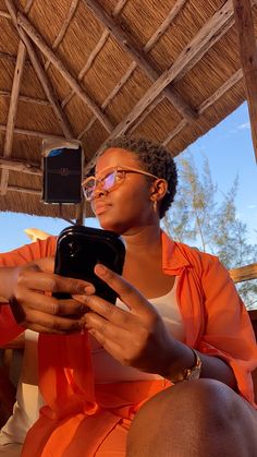 a woman in an orange dress is looking at her cell phone while sitting under a straw umbrella