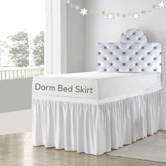 a white bed with a skirt on it in a room next to a window and rug