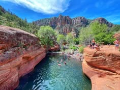 people are swimming in the water near red rocks and cliffs with green trees on either side