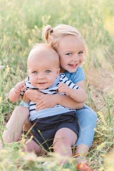 two young children sitting on the ground in tall grass, one is hugging the other