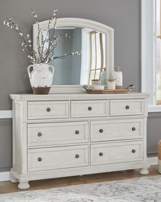 a white dresser with a mirror and vase on it's top, in front of a gray wall