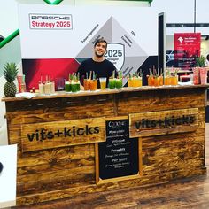 a man standing behind a wooden counter filled with juices and smoothie drinks in front of a sign