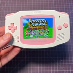 a hand holding a pink and white nintendo game boy advance with the words harvest moon on it