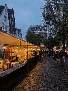 an outdoor market with people walking around and buildings in the background at dusk, on a cobblestone street