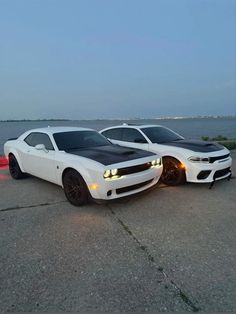 two white muscle cars parked next to each other in front of the ocean at dusk