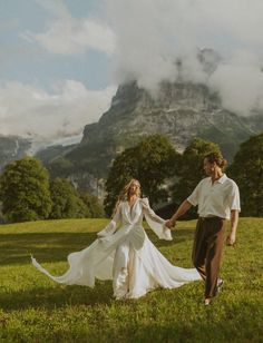 a bride and groom holding hands while walking through the grass with mountains in the background