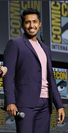 a man in a purple suit and pink shirt smiles as he stands next to a microphone