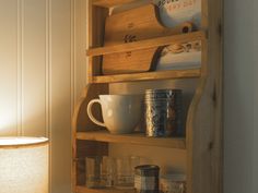 a wooden shelf with cups and mugs on it in front of a wall lamp