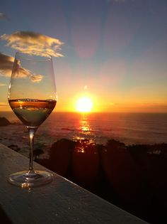 a glass of wine sitting on top of a wooden table next to the ocean at sunset