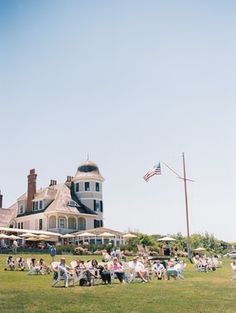 people are sitting on the grass in front of a large house with an american flag