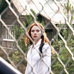 a woman standing behind a chain link fence in front of a house and looking at the camera