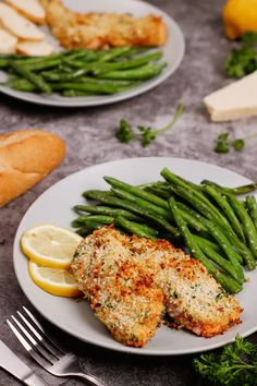 two white plates topped with fish and green beans next to breaded buns on a table