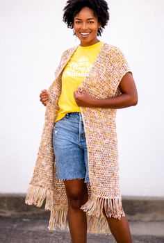 a woman standing in front of a white wall wearing a yellow t - shirt and fringed cardigan