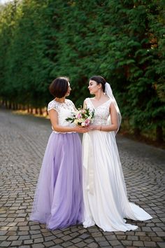 two women standing next to each other on a cobblestone road with trees in the background