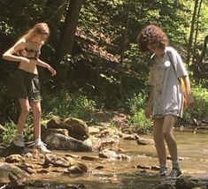 two young women standing on rocks in the water near some trees and rocks, while one looks at her cell phone